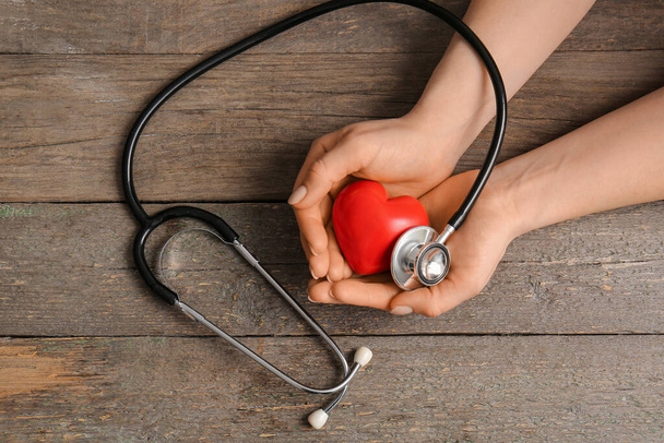 Mani femminili con cuore rosso e stetoscopio su sfondo di legno - Foto, immagini