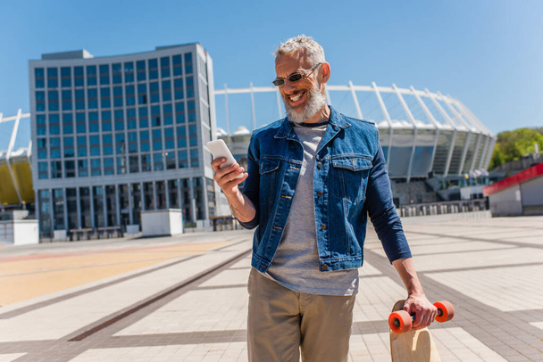 happy middle aged man in sunglasses holding longboard while using cellphone on urban street - Foto, imagen