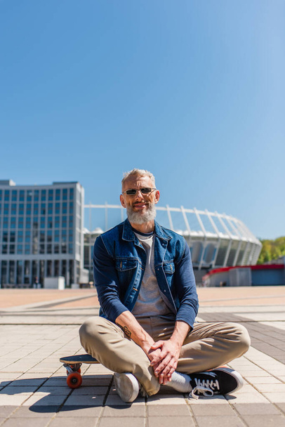 positiver Mann mittleren Alters mit Sonnenbrille sitzt draußen auf Longboard  - Foto, Bild