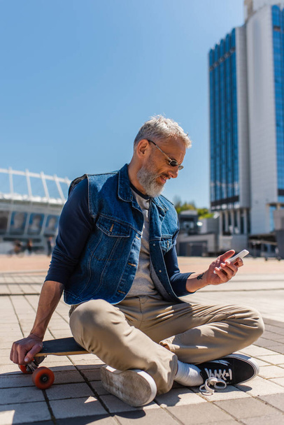 happy middle aged man in sunglasses sitting on longboard while using cellphone on urban street - Zdjęcie, obraz