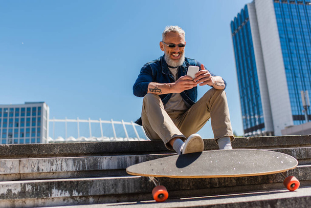 happy middle aged man in sunglasses sitting on stairs near longboard while using cellphone on urban street - Photo, Image
