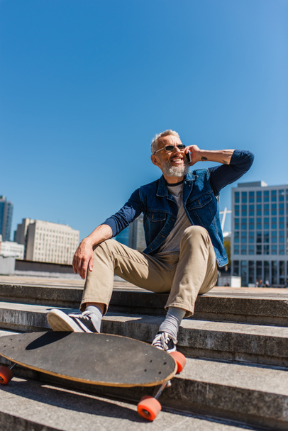 happy middle aged man in sunglasses sitting on stairs near longboard while talking on cellphone on urban street - Φωτογραφία, εικόνα
