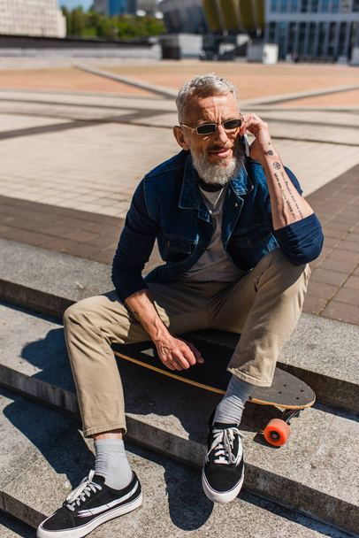 tattooed middle aged man in sunglasses sitting on stairs near longboard while talking on cellphone on urban street - Photo, image