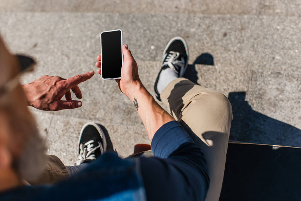 Draufsicht auf tätowierten reifen Mann zeigt auf Smartphone mit leerem Bildschirm in der Nähe von Longboard - Foto, Bild
