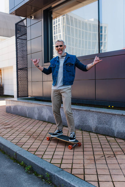 full length of excited man with open mouth riding longboard and showing rock sign on urban street - Zdjęcie, obraz