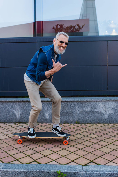 full length of happy man with open mouth riding longboard and showing rock sign on urban street - Foto, Imagem