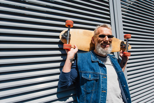bearded middle aged man in sunglasses smiling and holding longboard  - Фото, изображение