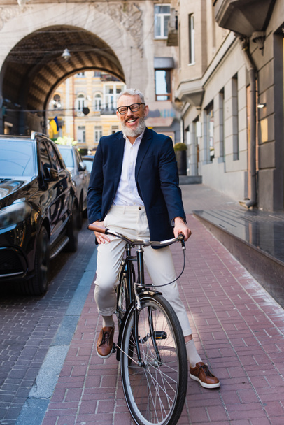 glücklicher bärtiger Mann mit Brille fährt Fahrrad auf der Stadtstraße  - Foto, Bild