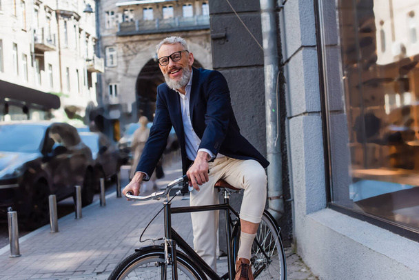 positive and bearded man in glasses riding bicycle on urban street  - Foto, immagini