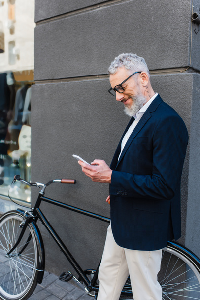 happy mature man in glasses using smartphone near bicycle on urban street  - Photo, Image