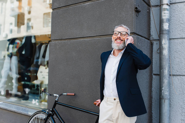happy middle aged man in glasses talking on smartphone near bike on urban street - Photo, image