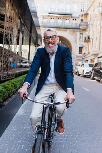 pleased middle aged man in glasses riding bicycle on modern urban street  - Foto, Imagem