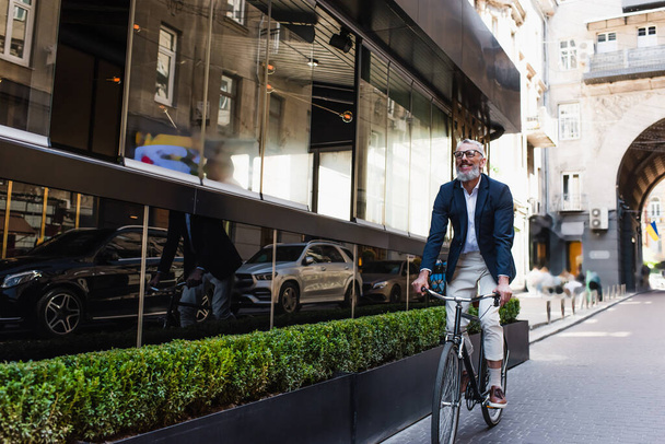 smiling middle aged man in blazer and glasses riding bicycle on modern urban street  - Photo, Image