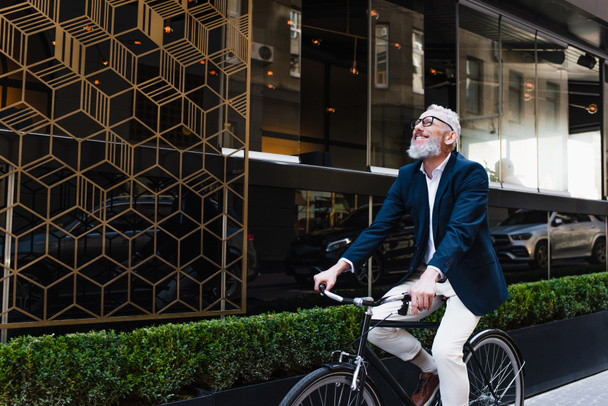 joyful middle aged man in blazer and glasses riding bicycle on modern urban street  - Zdjęcie, obraz