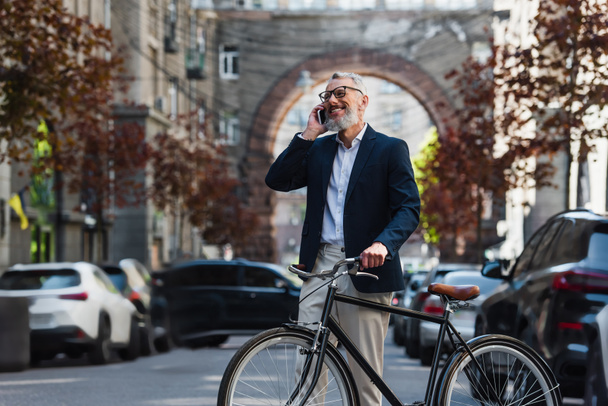 joyful middle aged man in blazer and glasses talking on smartphone and standing near bicycle on modern urban street  - Photo, Image