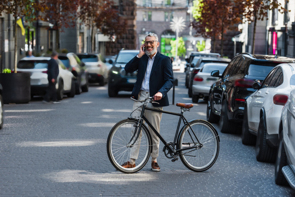 full length of happy middle aged man in blazer and glasses talking on smartphone and standing near bicycle on modern urban street  - Photo, Image