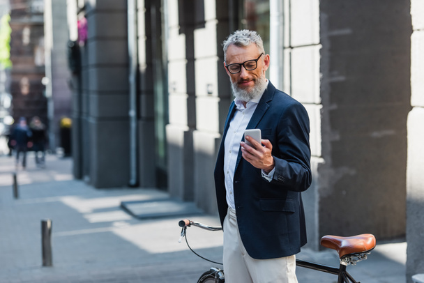 Mann mittleren Alters in Blazer und Brille schaut aufs Smartphone und steht neben Fahrrad auf der Straße  - Foto, Bild