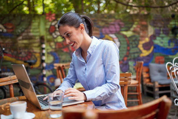 portrait of young beautiful caucasian adult female sitting in outdoor cafe, smiling, looking at her laptop, typing. online communication concept - Fotografie, Obrázek