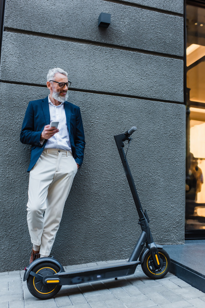cheerful mature businessman in blazer standing near e-scooter and using smartphone  - Zdjęcie, obraz