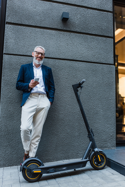 smiling mature businessman in blazer standing near e-scooter and using smartphone  - Foto, imagen