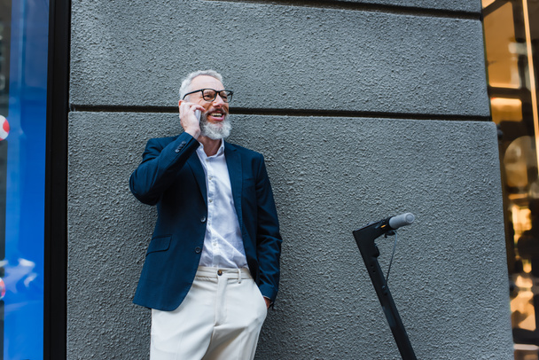 happy mature businessman in blazer talking on cellphone near e-scooter  - Photo, image