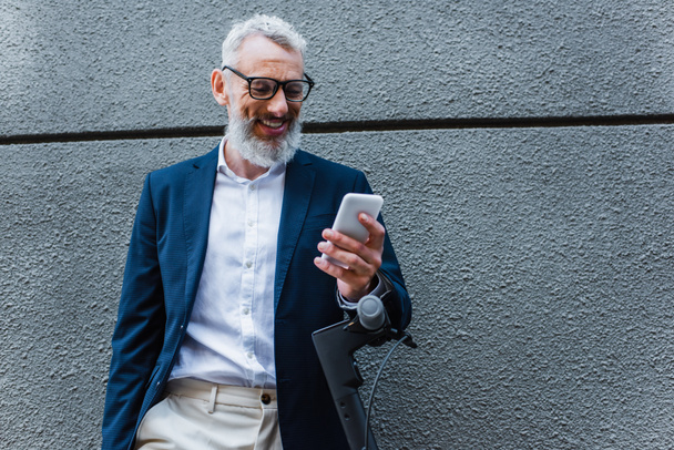 улыбающийся взрослый бизнесмен в блейзере с помощью смартфона возле электронного скутера - Фото, изображение