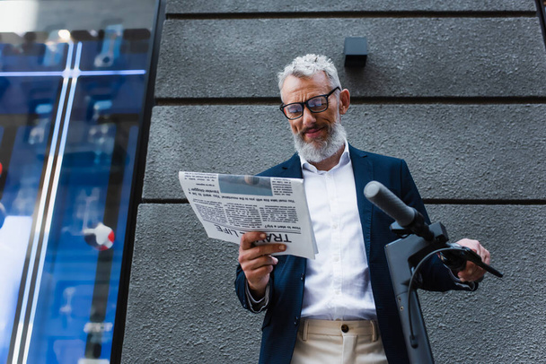 Низкий угол обзора счастливого зрелого бизнесмена в блейзере, читающего газету возле е-скутера  - Фото, изображение