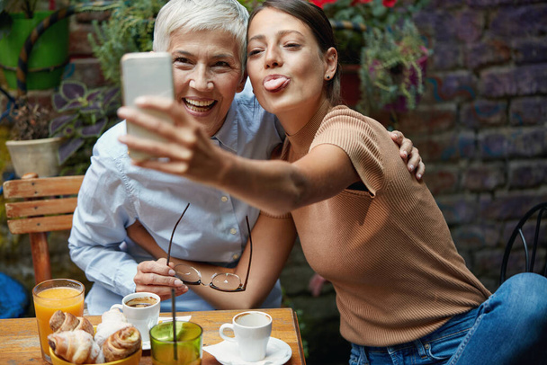A young girl is making funny faces while taking a selfie with her grandma in a pleasant atmosphere at a bar. Leisure, bar, friendship, outdoor - Photo, image