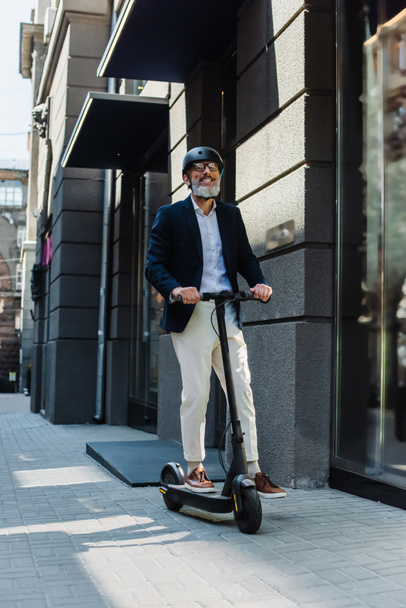 full length of happy mature businessman in blazer and helmet riding e-scooter  - Valokuva, kuva