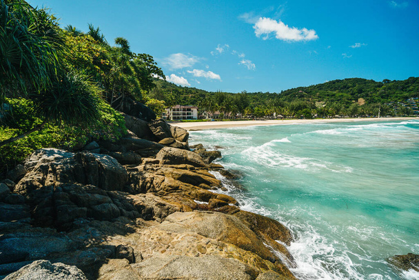 Скалистый берег с зелеными кустами на фоне тропического пляжа с некоторыми зданиями и зелеными холмами. Летние каникулы и приключения на природе.. - Фото, изображение