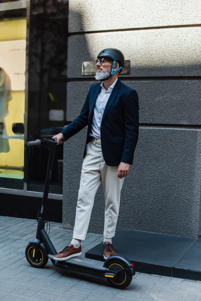 full length of mature businessman in blazer and helmet standing near e-scooter  - Foto, Imagem
