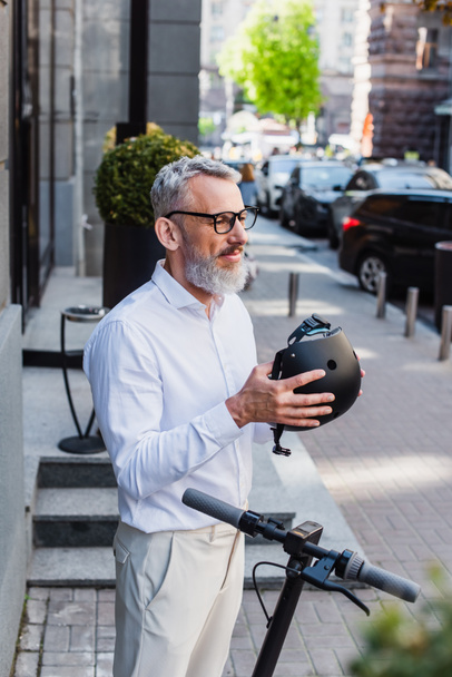 smiling and mature man in shirt holding helmet near electric scooter - Фото, изображение