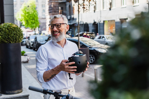 smiling and mature man in shirt holding helmet near electric scooter on street  - Fotografie, Obrázek
