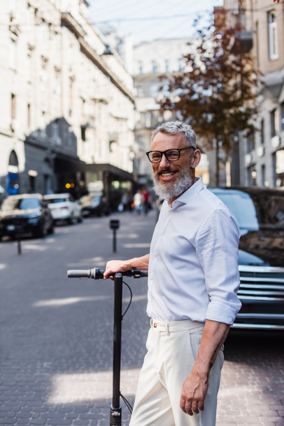 smiling middle aged man in white shirt near electric scooter on street  - Photo, Image