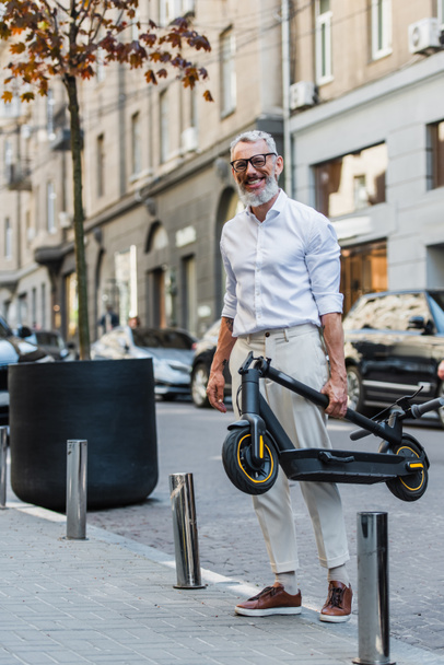 smiling middle aged man in white shirt carrying electric scooter on street  - Fotoğraf, Görsel