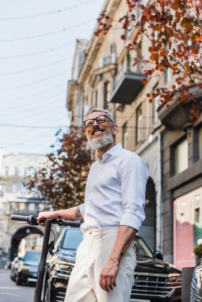 low angle view of joyful middle aged man in white shirt looking up near electric scooter on street  - Fotografie, Obrázek