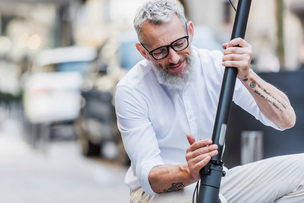 Lächelnder und reifer Mann mit Brille stellt Elektroroller auf der Straße ein  - Foto, Bild