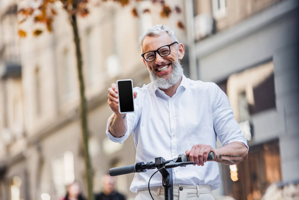 happy middle aged man in glasses holding cellphone with blank screen and standing near e-scooter  - 写真・画像