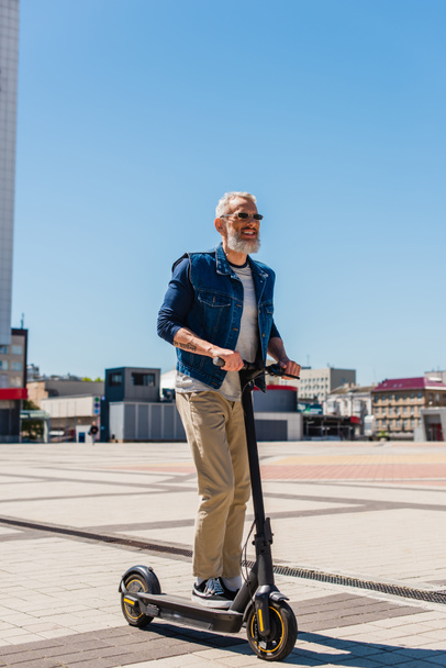 full length of cheerful man in sunglasses riding electric scooter in urban city  - Foto, afbeelding