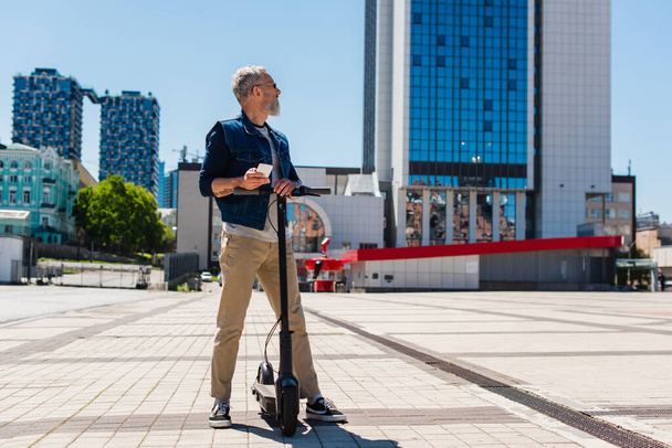 Vollbärtiger Mann mit Sonnenbrille benutzt Handy in der Nähe von Elektroroller auf Stadtstraße - Foto, Bild