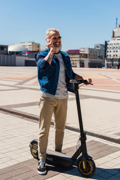 full length of cheerful man in sunglasses talking on cellphone near electric scooter on urban street - Valokuva, kuva