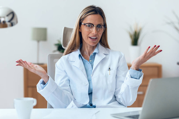 Portrait of mature attractive female doctor looking at camera while working sitting in the consultation. - Photo, Image