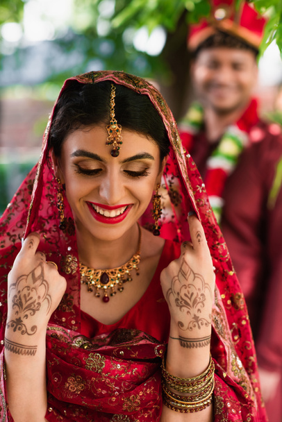 cheerful indian bride in sari and headscarf near blurred man in turban on background  - Фото, изображение