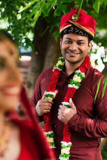gelukkig indiaanse man in tulband en bloemenslinger kijken naar wazig bruid  - Foto, afbeelding