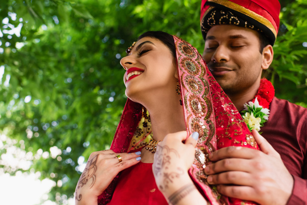 низкий угол зрения довольный индийский мужчина в тюрбане обнимая невесту в красном сари  - Фото, изображение