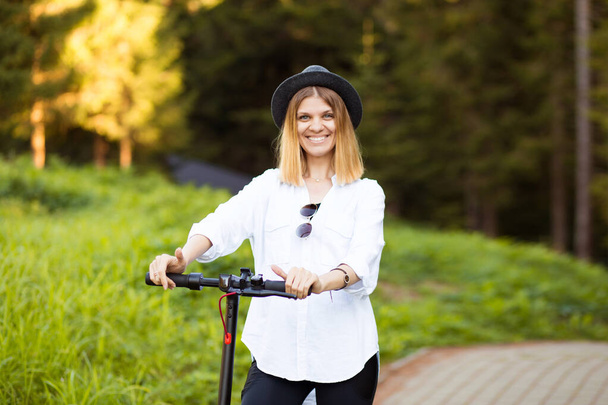 Μοντέρνα ξέγνοιαστη γυναίκα σε λευκό πουκάμισο και μαύρο καπέλο βόλτα με το σκούτερ λάκτισμα εξωτερική στο δρόμο καλοκαιρινό πάρκο. Ηλεκτρικές οικολογικές μεταφορές. - Φωτογραφία, εικόνα