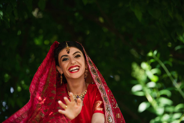 happy indian bride in red sari and traditional headscarf with ornament gesturing while looking at camera - Photo, Image