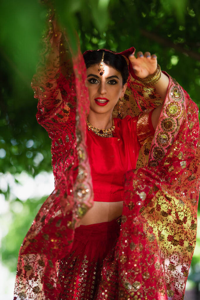 young indian bride in red sari and traditional headscarf with ornament posing outside  - Фото, зображення