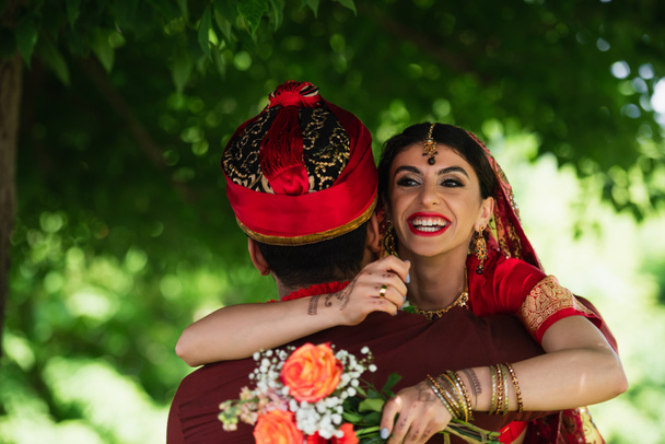 back view of indian man in turban hugging cheerful bride in traditional headscarf holding flowers  - Foto, Imagen