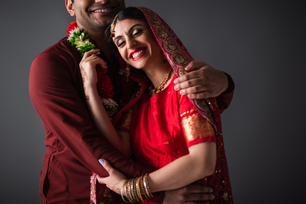 Indian man in turban hugging happy bride in traditional headscarf isolated on grey  - Photo, Image